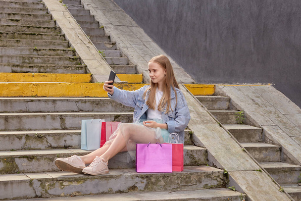 Cute girl takes a selfie sitting on the stairs with shopping in colorful bags. - 写真・画像