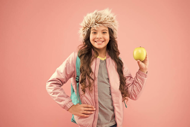 small happy girl earflap hat eat apple. end of winter semester. holiday and vacation. kid pink backdrop. teen daily life. education and happy people schoolgirl with books after lesson. school break - Foto, immagini