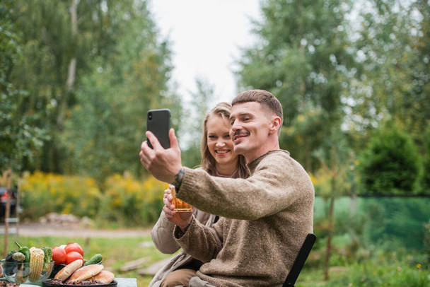 Una giovane bella coppia scattare foto della loro nuova casa di campagna su una fotocamera smartphone. Picnic nella natura in occasione dell'acquisto di immobili in campagna. - Foto, immagini