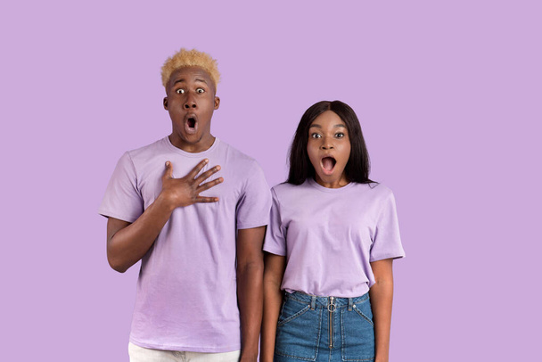 Choqué couple afro-américain ouvrir la bouche en état de choc, mettre la main sur le cœur sur fond violet studio - Photo, image