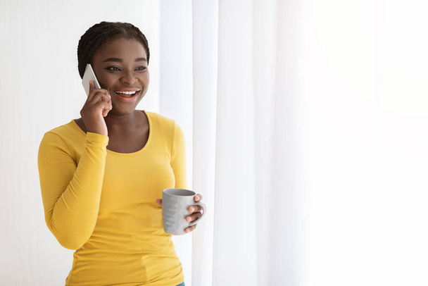 Opgewonden zwarte vrouw met mobiele telefoon en koffie in de buurt van venster Genieten van telefoon bellen - Foto, afbeelding