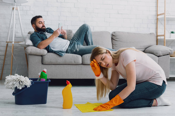 Женщина страдает от головной боли и делает уборку дома, мужчина с бородой лежит на диване и играет по телефону - Фото, изображение