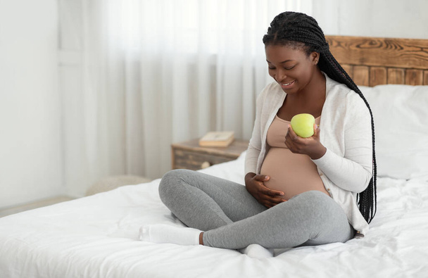 Dieta saludable para el embarazo. Embarazada africana comiendo manzana mientras descansa en la cama - Foto, imagen