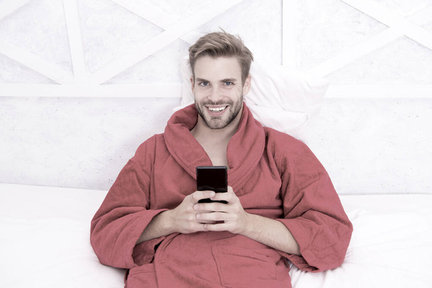 Living a mobile lifestyle. Cheerful guy using mobile device in bed. Handsome man smiling with mobile phone in hands. Mobile communication and wireless technologies in home - Foto, Bild
