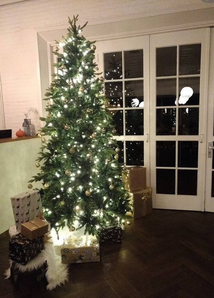 Árbol de Navidad con regalos se enciende en un acogedor apartamento por la noche. Navidad y Año Nuevo decoración del hogar - Foto, imagen