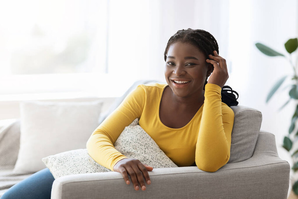 Позитивна афроамериканська леді позує в домашньому інтер'єрі, сидячи на зручному дивані
 - Фото, зображення