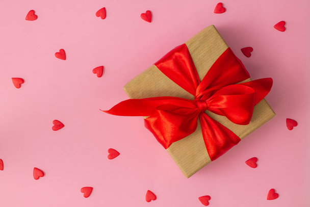 Gift wrapped in craft paper with red ribbon and bow and red hearts on pink background. Flat lay. Copy space. - Valokuva, kuva