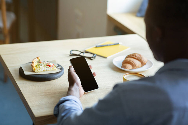 Gros plan de l'homme noir tenant téléphone portable avec écran vide tout en déjeunant au café, maquette pour la conception d'applications mobiles - Photo, image