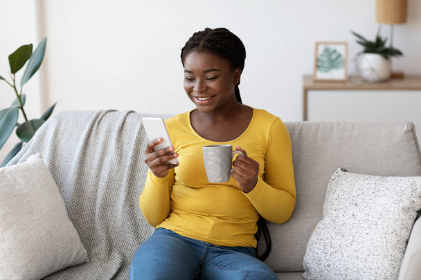 Jeune femme noire avec smartphone et café relaxant dans le salon - Photo, image