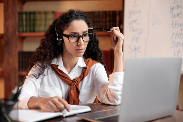Konzentrierte Frau sitzt am Schreibtisch, trägt Headset, benutzt Computer - Foto, Bild