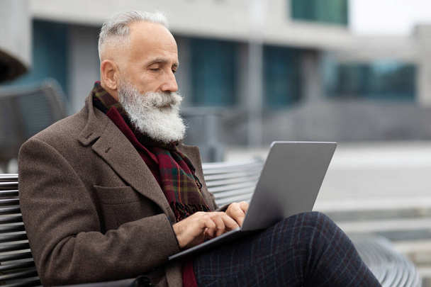 Homem idoso de cabelos grisalhos concentrado sentado ao lado do prédio de escritórios, usando laptop - Foto, Imagem