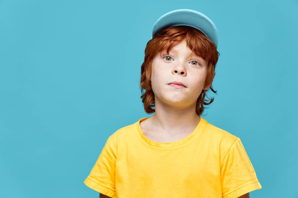 Roodharige jongen met een verrassende uitdrukking op zijn gezicht blauwe pet gele t-shirt blauwe achtergrond  - Foto, afbeelding