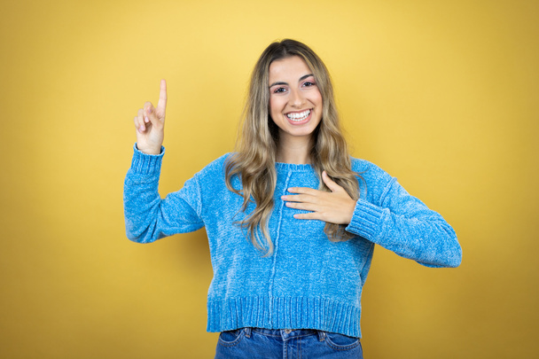 Pretty blonde woman with long hair standing over yellow background smiling swearing with hand on chest and fingers up, making a loyalty promise oath - Photo, Image