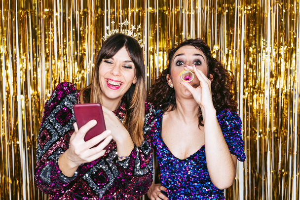 Two beautiful women with party clothes and Christmas headbands having fun together in a New Year's Eve party with some portraits of them. New Year's Eve concept - Photo, Image