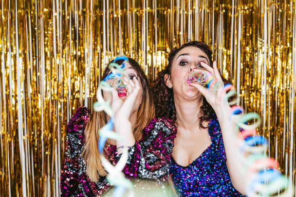 Twee mooie vrouwen in elegante kleren met kerstversieringen achter hen blazen slingers naar de camera terwijl vieren in een nieuwjaarsfeest. Nieuwjaarsfeest concept - Foto, afbeelding