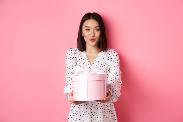Cute asian girlfriend congratulate with valentines day, giving cute romantic gift in box, pucker lips for kiss, standing over pink background - Foto, Imagem