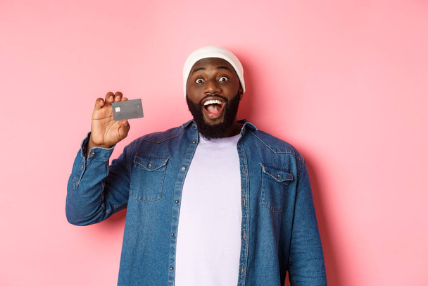 Σχέδιο αγορών. Εντυπωσιασμένος μαύρος τύπος που δείχνει πιστωτική κάρτα, κοιτάζει την κάμερα εντυπωσιασμένος, συστήνει τράπεζα, στέκεται πάνω από ροζ φόντο - Φωτογραφία, εικόνα