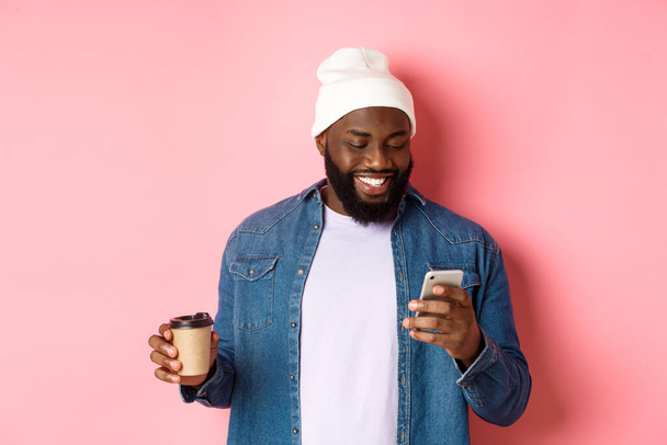 Wizerunek stylowego czarnego człowieka hipstera pijącego kawę na wynos, czytającego wiadomość przez telefon i uśmiechniętego, stojącego nad różowym tłem - Zdjęcie, obraz