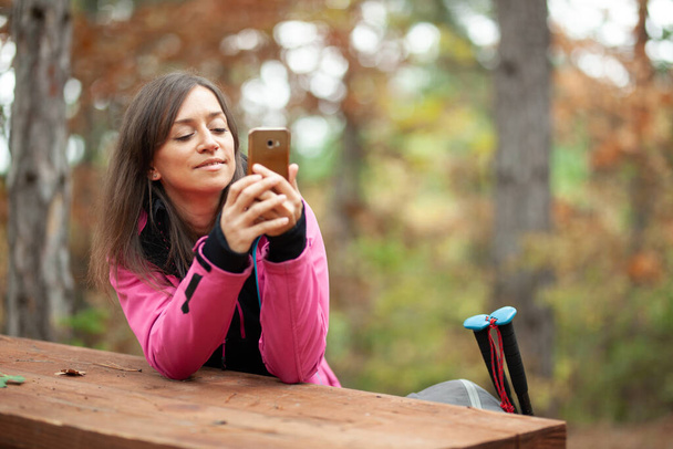 Hiker girl resting on a bench in the forest. Backpacker with pink jacket holding cell phone. - Foto, Imagem
