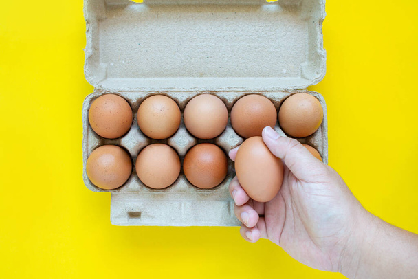 Man's hand is picking up a brown egg in a carton box. On a yellow background. - Fotoğraf, Görsel