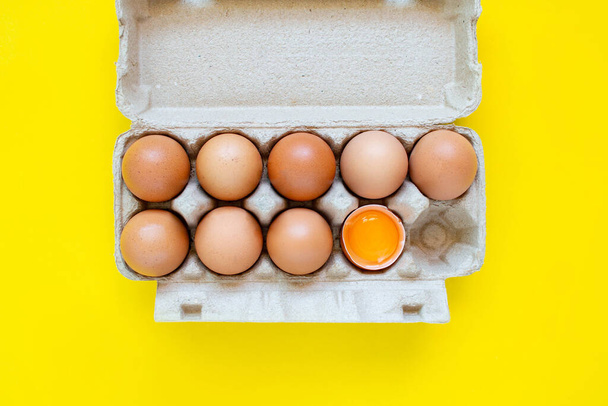 Primeros planos huevos marrones agrietados y huevos en cajas de papel colocados uno al lado del otro sobre un fondo amarillo. - Foto, Imagen