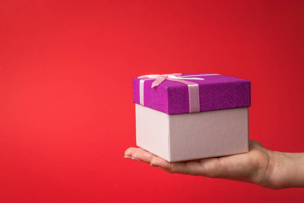 A woman's hand with a gift box on a bright red background. Surprise in the hands of a girl. - Foto, Imagen