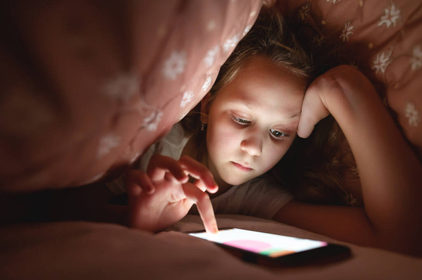 Young girl ying under the covers. playing with mobile phones and checking email in the bedroom in the apartment. Childrens gadget addiction concept - Φωτογραφία, εικόνα