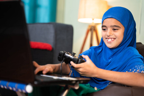 Happy smiling Muslim Girl Kid gioca online con il videogame sul laptop utilizzando gamepad o joystick a casa indossando un vestito hijab - concetto di bambino che utilizza la tecnologia e lo stile di vita moderno - Foto, immagini
