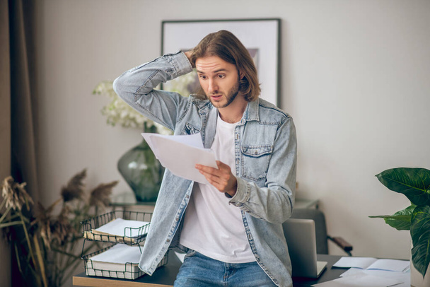 Young man in jeans shirt holding papers and looking surprised - Foto, afbeelding