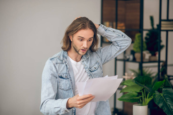Young man in jeans shirt holding papers and looking surprised - Foto, immagini