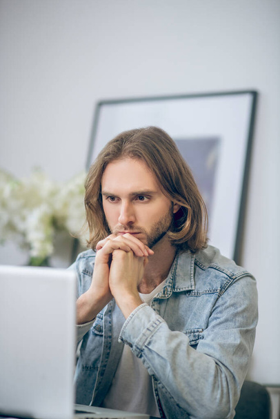 Long-haired man in jeans shirt working on laptop and looking thoughtful - Φωτογραφία, εικόνα