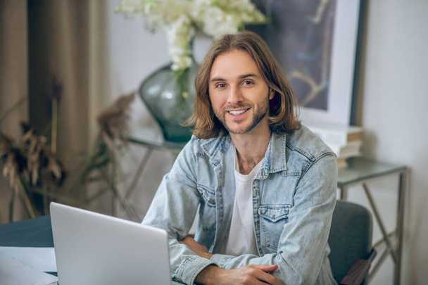 Long-haired man in jeans shirt working on laptop and looking happy - Foto, Imagem