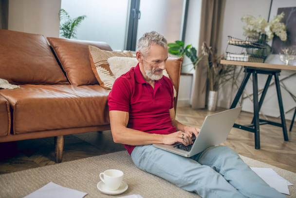 Grey-haired man sitting on the floor and typing on a laptop - 写真・画像
