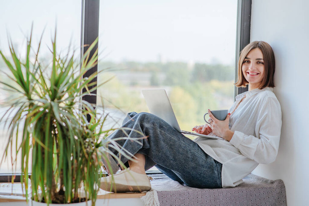 Gyönyörű barna fiatal nő alkalmi ruhák ül az ablak mellett a laptop a munka vagy az online oktatás otthonról. Társadalmi távolság az e-tanulás vagy az otthoni koncepciótól való munkavégzés terén. - Fotó, kép