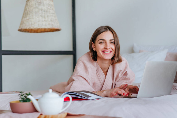 Young smiling woman in bath robe lying on the bed using laptop for work or messaging. Morning blogger routine concept. - Photo, image