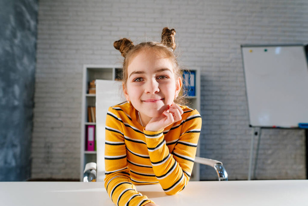 Szczęśliwy uśmiech 8 lat dziewczyna powiedzieć cześć patrząc na aparat siedzi przy biurku w pokoju dzieci podczas lekcji online. - Zdjęcie, obraz