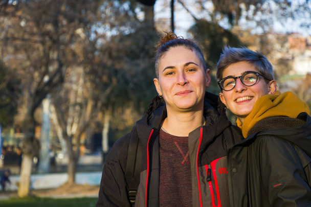 Портрет пары геев-лесбиянок, две взрослые женщины в парке, улыбающиеся и обнимающиеся, время заката - Фото, изображение