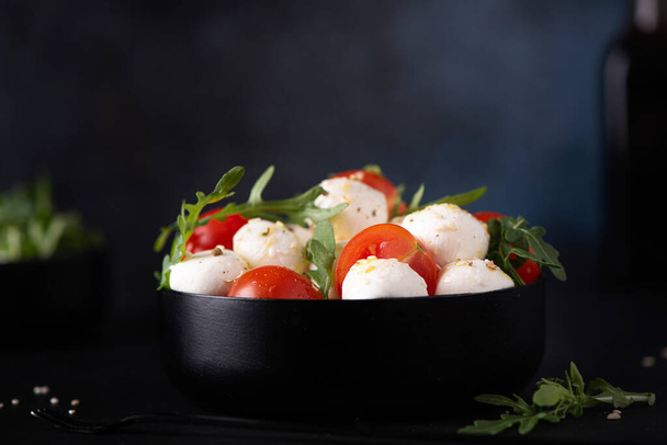 mozzarella salad of cherry tomatoes and arugula seasoned with olive oil in a black bowl, close up - Foto, imagen
