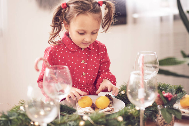 Söpö kihara lapsi tyttö seisoo joulupäivällispöydässä asettumassa ruokia valmistautuu juhlimaan jouluaattoa koristeltu ruokasali, jossa on puu ja valot. - Valokuva, kuva