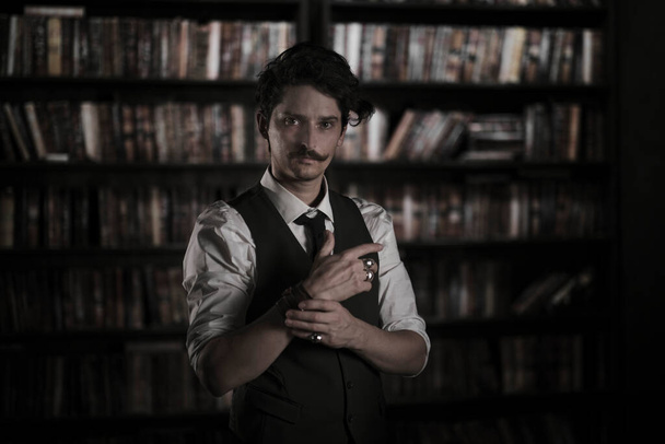 暗い図書館の部屋で男は夜に立っている。口髭を生やしたスタイリッシュな男がブレスレットをまっすぐにし. - 写真・画像