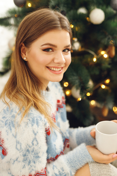Side view of girl wears festive clothes take cup of coffee at home near Christmas Tree. Smiling woman sit on mat in blue sweater with deer and gray stockings - Foto, Imagen