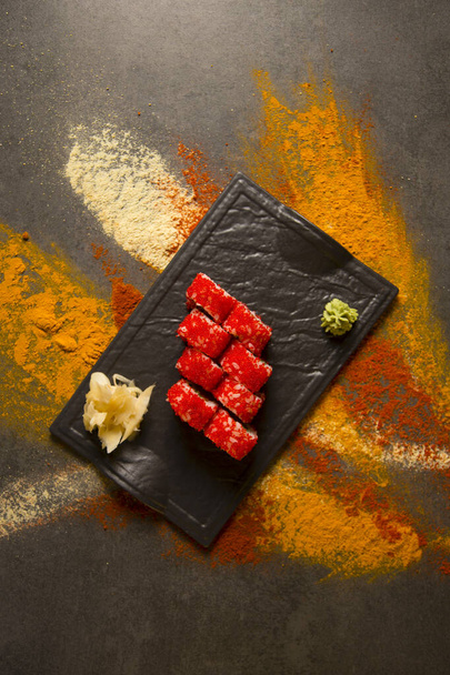 Direkt über der japanischen Sushi-Rolle mit rotem fliegendem Fischrogen Tobiko serviert mit Wasabi und Ingwer auf schwarzem Teller. Kurkuma-Pulver, trockener Knoblauch, Paprika-Gewürze auf dunklem Hintergrund. Food-Art-Konzept - Foto, Bild