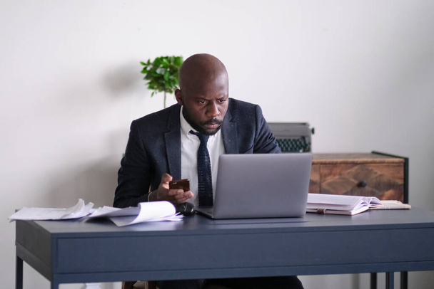 Un jeune dirigeant africain utilise un smartphone, lit de mauvaises nouvelles et travaille sur un ordinateur portable alors qu'il est assis à son bureau dans un bureau. foyer doux - Photo, image