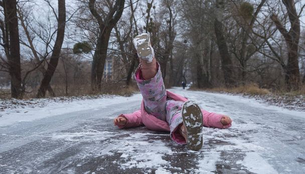 Kind meisje ligt op de weg, neergang en ongeluk in het winterseizoen, zwart ijs. Risico van ongevallen in de winter - Een vrouw gleed uit op een gladde sneeuwweg. - Foto, afbeelding
