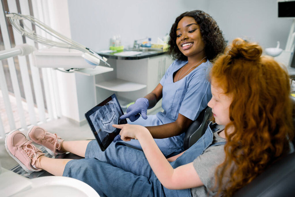 Little patient kid girl with curly red hair sitting in dentistry chair and pointing on the tablet with teeth panoramic radiography. Pretty african woman doctor explaining the ways of treatment - Foto, imagen