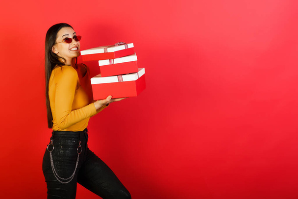 Mujer joven sostiene pesadas cajas de regalo grandes y de pie cerca de fondo rojo con espacio para la publicidad. Concepto de vacaciones y regalos. - Foto, imagen