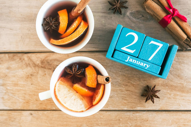 27 Ιανουαρίου. Μπλε κύβος ημερολόγιο με μήνα και ημερομηνία και φλιτζάνια με ζεστό κρασί, ξύλινο φόντο. - Φωτογραφία, εικόνα