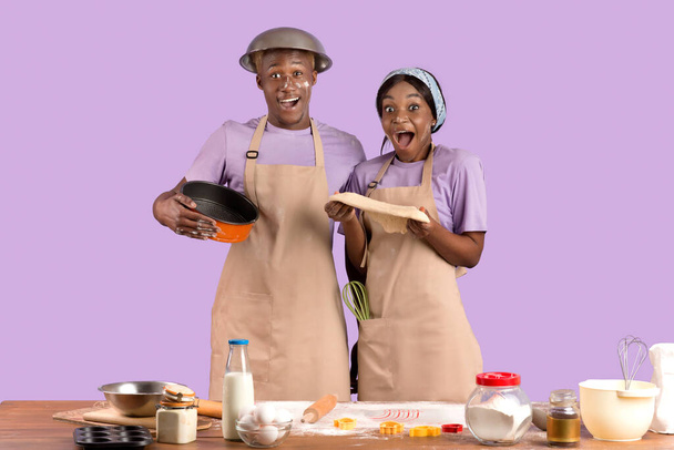 Взволнованная черная пара готовит вместе, держа домашнее тесто на фоне фиолетовой студии - Фото, изображение