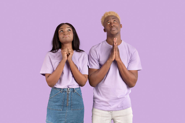 Religieus zwart paar die handen bij elkaar steken in gebed, hun ogen opheffen, God om hulp vragen op een lila achtergrond - Foto, afbeelding