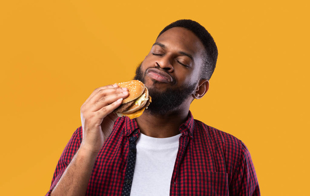 Αφρικανική Millennial Guy μυρίζοντας Burger στέκεται σε κίτρινο φόντο στούντιο - Φωτογραφία, εικόνα